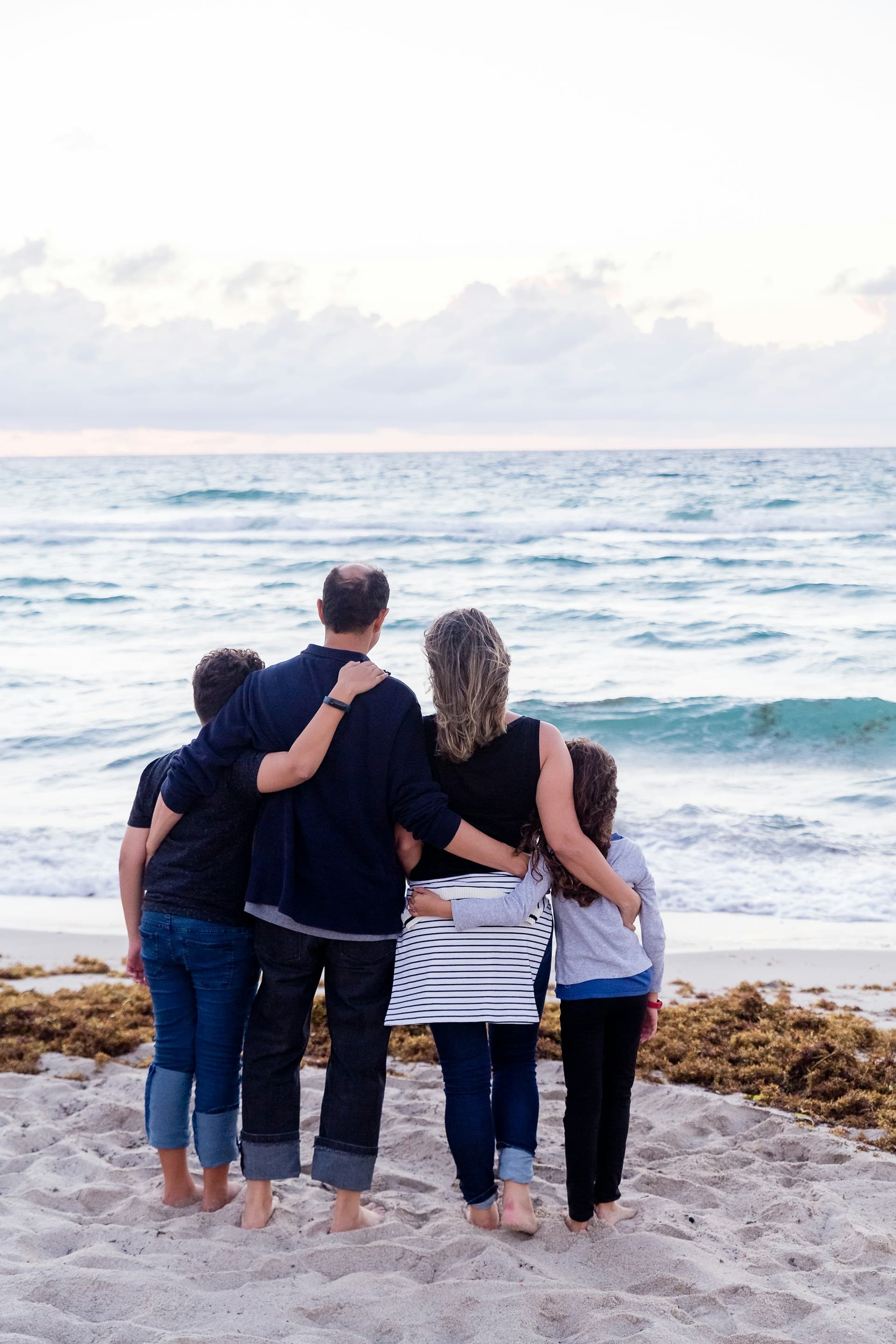 Family of four standing arm in arm on a sandy beach, looking out at the sea at dusk