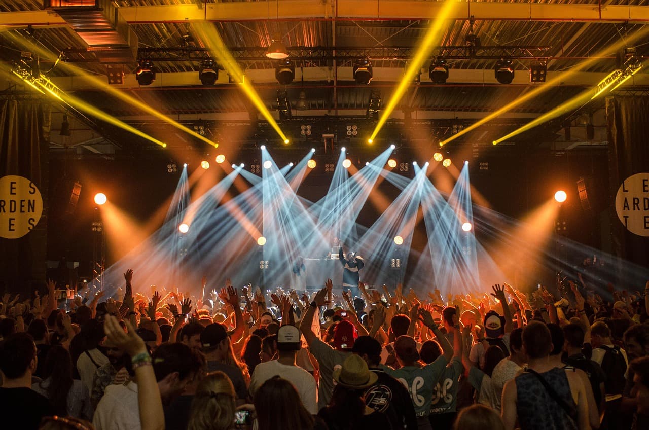 Crowd with hands raised at an indoor music event, lit by yellow and white stage beams