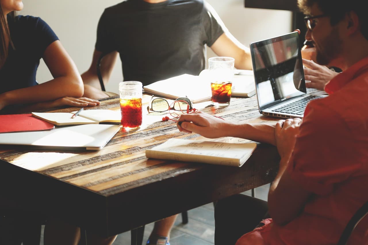 Team meeting around a wooden table with laptops, notebooks and drinks in a sunlit office